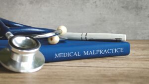 A blue medical law book titled "MEDICAL MALPRACTICE" on a wooden desk with a stethoscope and white pen for legal case documentation.