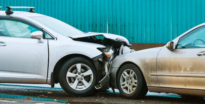 A silver sedan with roof rails has its entire front hood crumpled upward and the passenger-side headlight assembly destroyed where it has been driven into the rear quarter panel of a dusty gold-toned vehicle on a wet gray road surface with scattered debris visible on the pavement between the two interlocked cars and a corrugated teal metal wall running horizontally across the background — a close-up street-level view of the structural devastation caused by a serious car accident in Augusta GA.