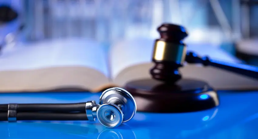 A dark black stethoscope with a ridged silver and blue chest piece lies coiled on a vivid blue reflective surface in the foreground with a large open hardcover book with yellowed pages spread flat in the center background and a dark mahogany judge's gavel with a gold band resting on its circular sound block to the right with a softly blurred blue-tinted legal setting- at Horst Shewmaker- visible behind all three objects — symbolizing the critical intersection of medical evidence and legal documentation at the core of every car accident injury case in Augusta GA.